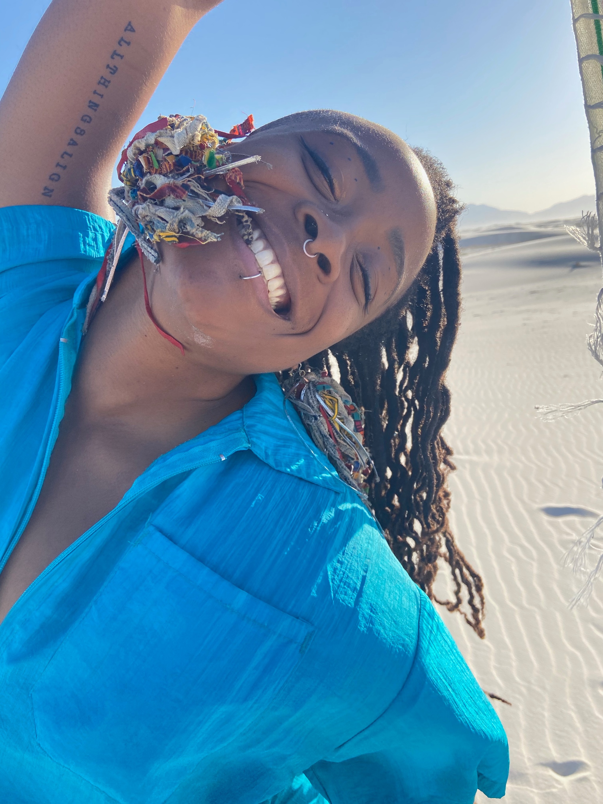 A smiling Black woman in a bright blue shirt leans over with with her head slightly back, one are raised, and a desert landscape in the background. She has a lip ring, nose ring, and a tattoo on the underside of her upper arm. 