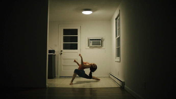 A dancer poses leaning forward on one bent knee in a dramatically lit kitchen