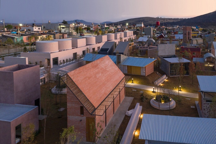 An overhead view of a residential area at dusk where unsual, contemporary housing meets a more traditional and modern neighborhood. In the foreground, one building made with pinkish brick looks almost like a traditional two-story house form, cut in two. Another series of cream-colored buildings employs cylindrical structures as a second story. Low hilltops rise in the distance to meet a peach and gray sky.