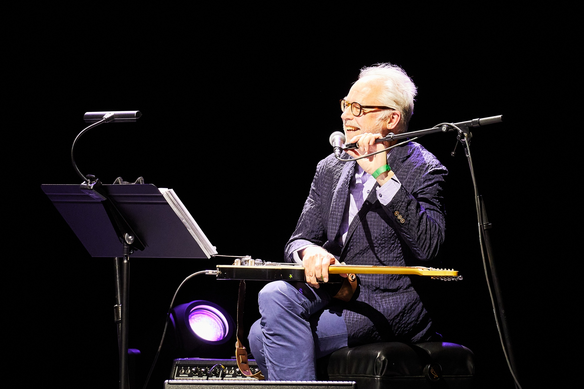 A guitarist seated on stage speaking into a microphone that he steadies in front of his face. He is a white man with white hair, wearing glasses and a blue blazer.