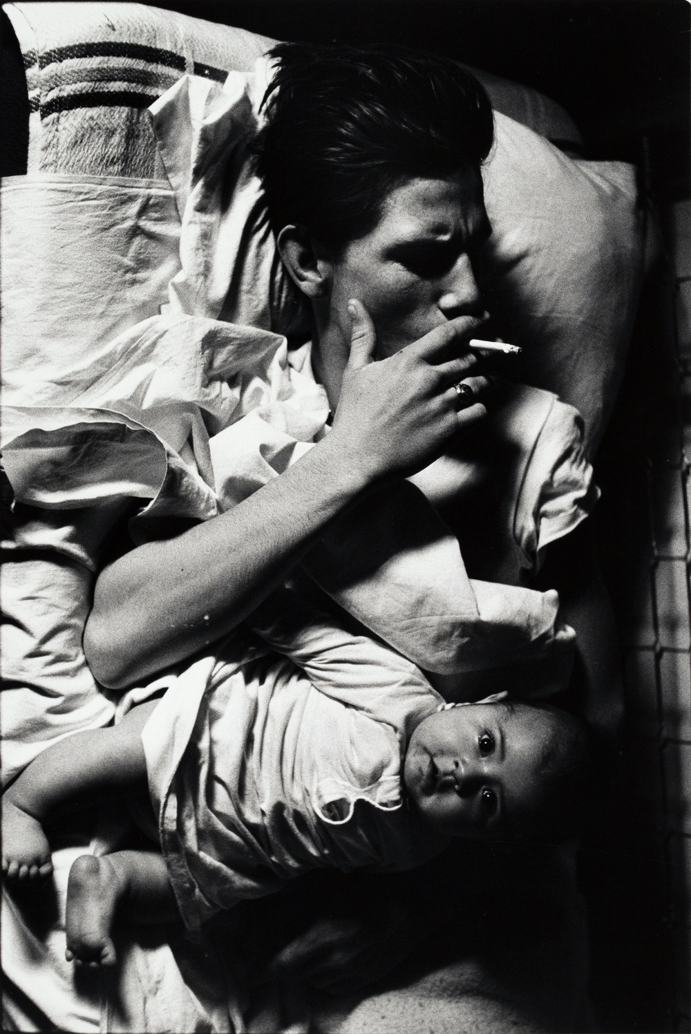 A black and white photograph looking down onto a man laying in bed smoking a cigarette with a baby laying on top of his torso who is looking directly up at the camera.