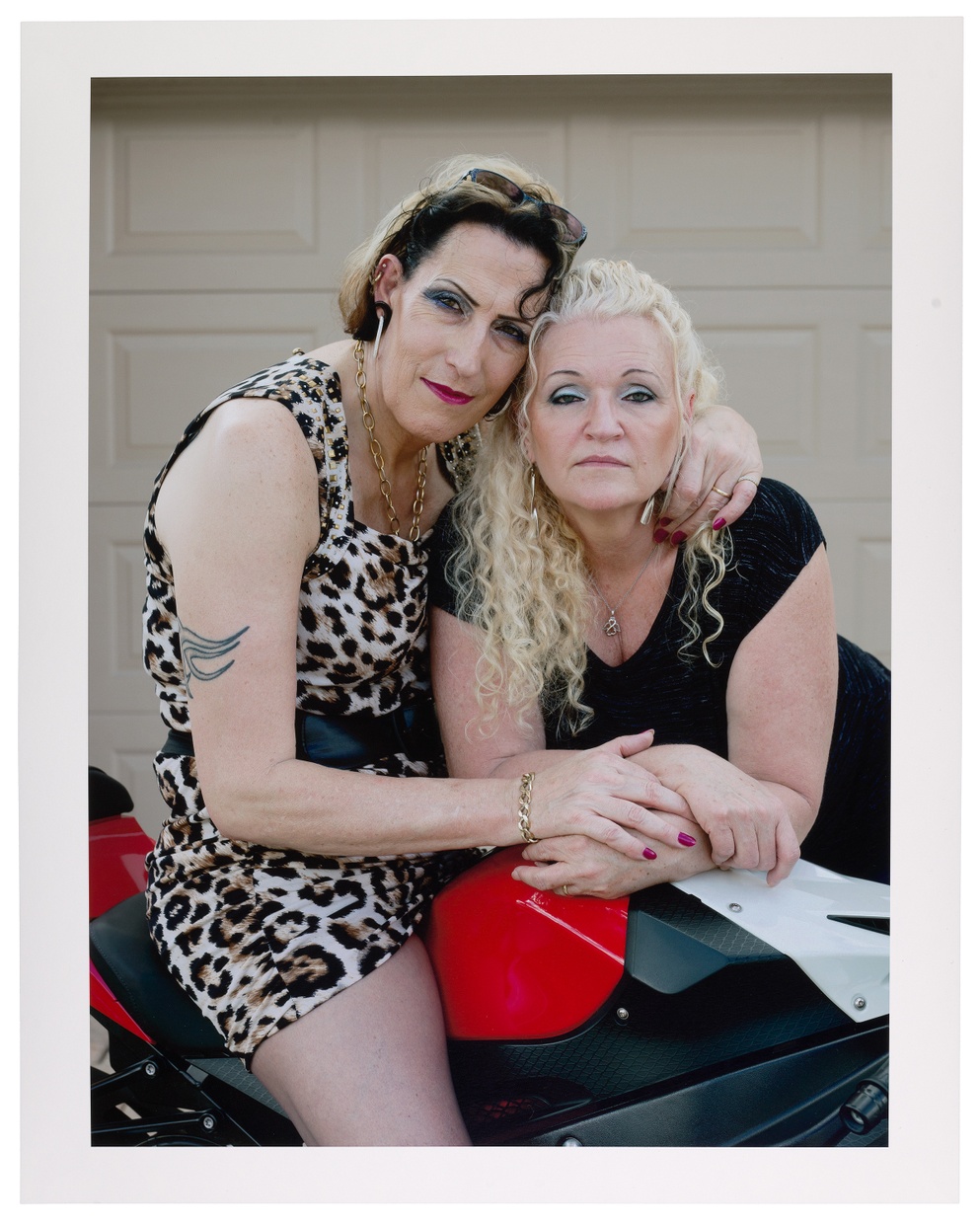 A photograph of two light-skinned people wearing dresses and heavy eye makeup embracing each other and leaning over a red and black motorcycle. 
