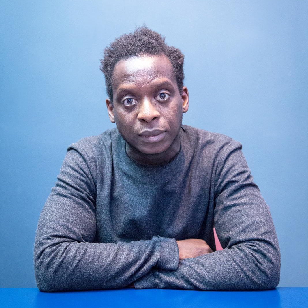 A portrait of Kobna Holdbrook-Smith, a Black actor who sits against a blue background with his arms folded on a blue table in front of him. He has short hair with a part down the left side of his head. He wears a grayish blue long sleeve shirt and looks intently at us with eyebrows raised.