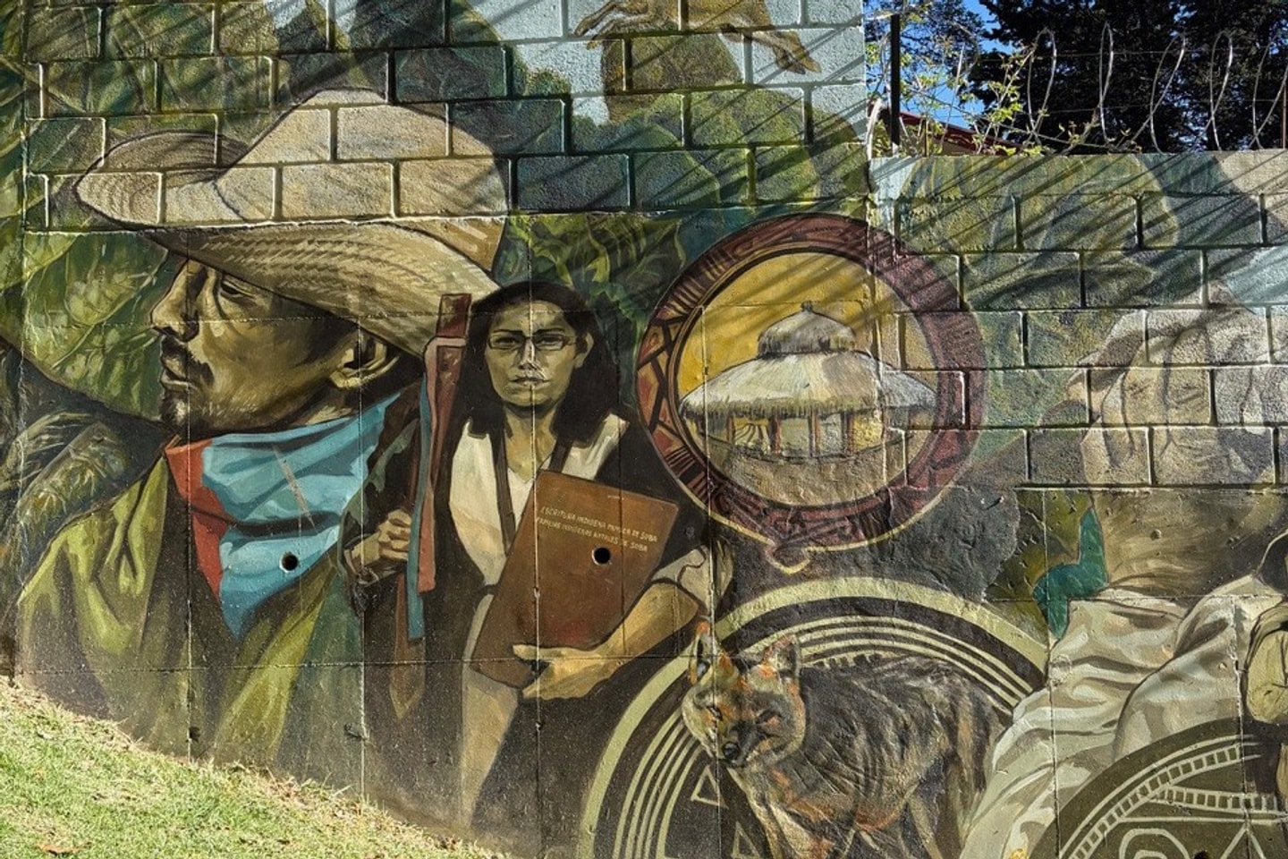 A colorful outdoor mural painted on a cinder-block wall topped with razor wire, set above a green lawn. The artwork depicts a person in a wide-brimmed straw hat and blue neckerchief, a figure holding an inscribed brown plaque, circular emblems featuring a thatched-roof building and a wolf, and traditional motifs.