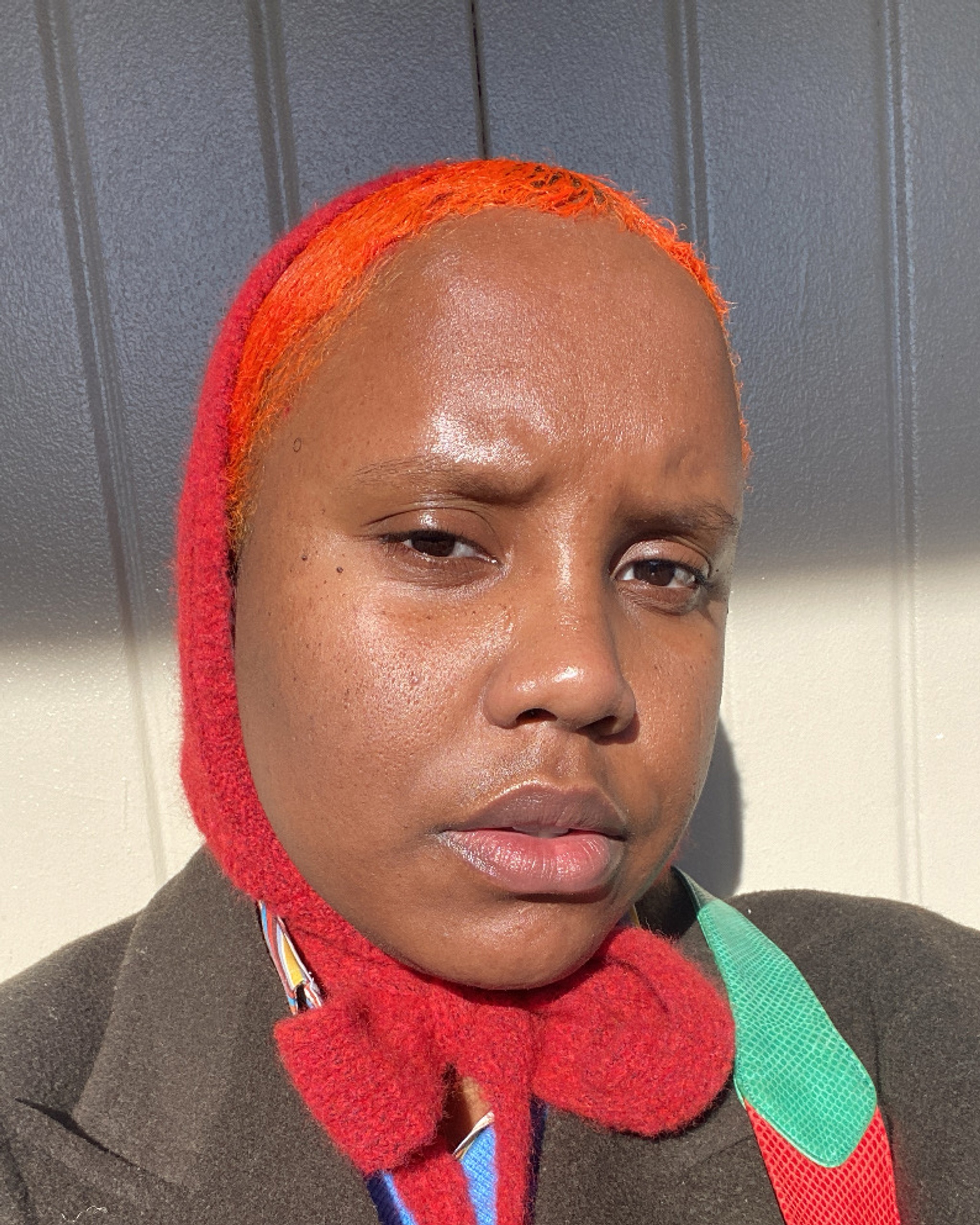 A portrait of Shala Miller, a Black nonbinary singer who is seen close up as if taking a selfie. Shala has a quizzical or skeptical expression. Their short hair is a bright orange and they wear a bright red scarf over their head, tied under their chin. 