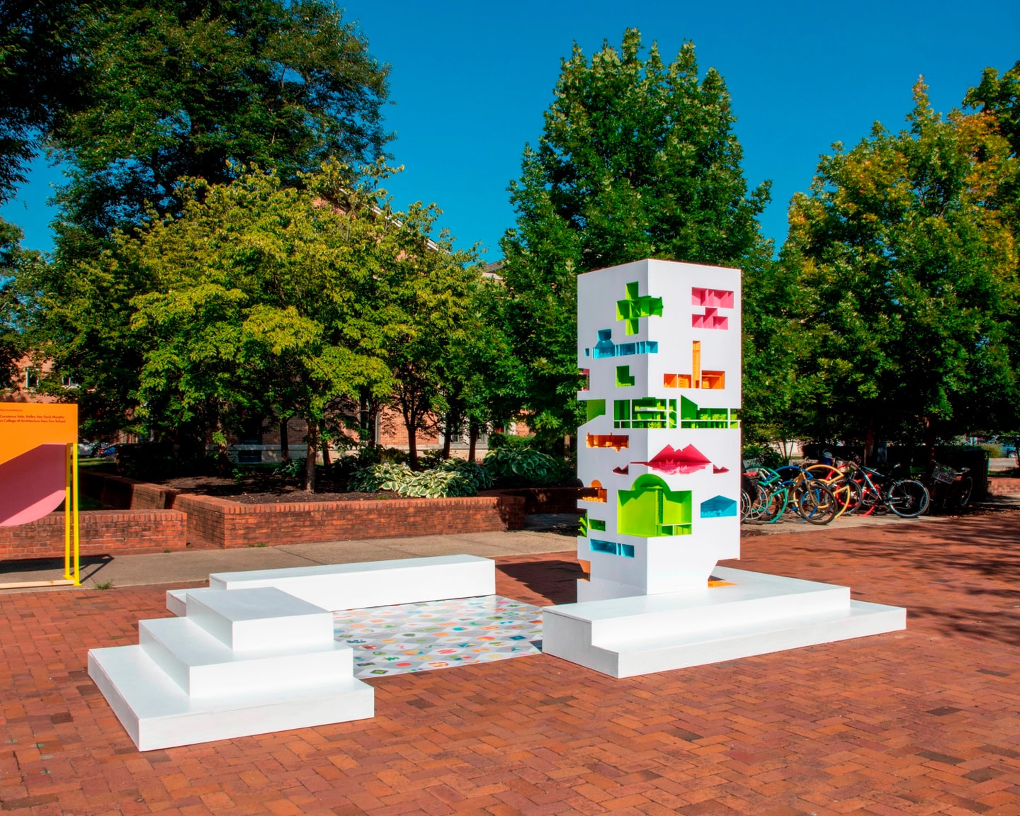 large white dollhouse with colorful sections cut into it with dolls made by students sitting outside in a plaza on brick with surroundings trees