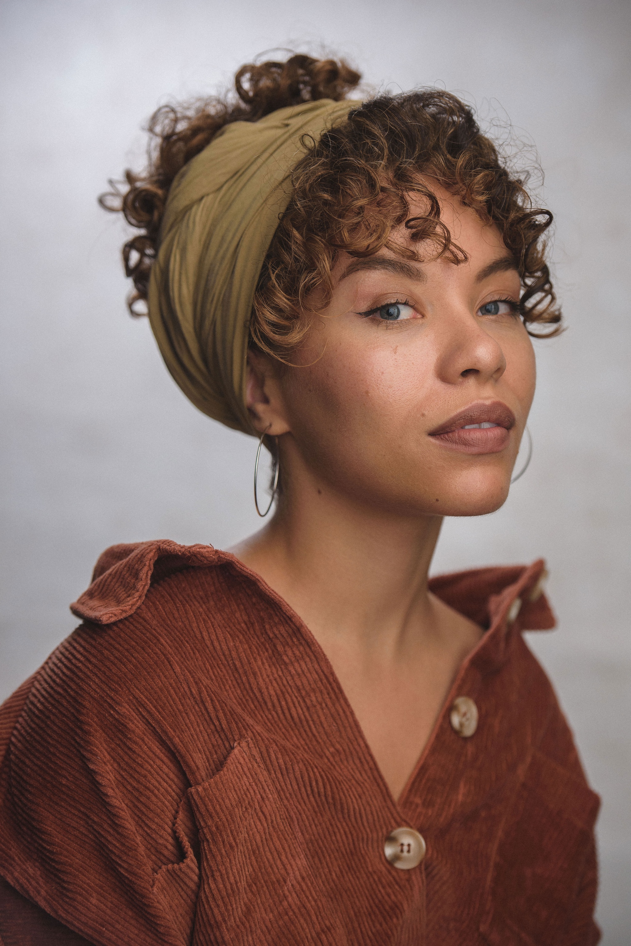 Saffron Coomber, a Black woman with curly brown hair pulled up in a wrap behind her head, poses against a neutral backdrop in three quarters view, turning her eyes to look directly at us. She wears a brown collared corduroy shirt.