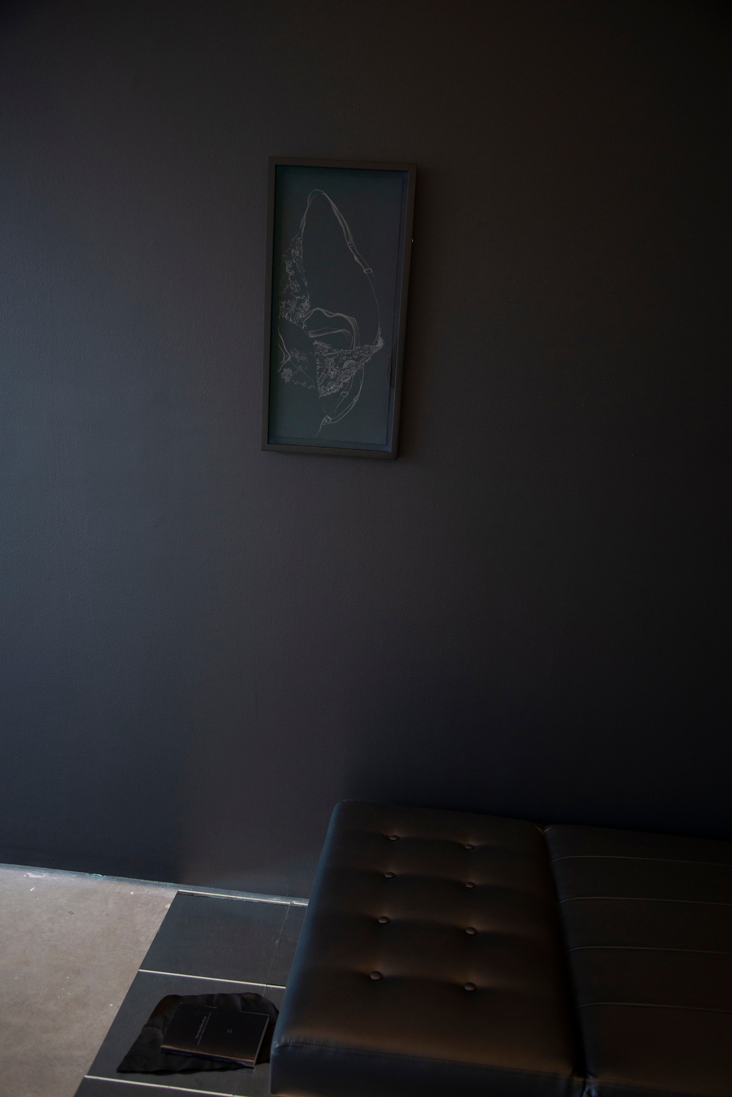 Against a black wall, on a black background in a tall, rectangular black frame, faint white lines that may resemble some kind of clothing or detail. Against the wall is a black bench on a light-hued floor with some reflective, possibly metal panels. On one of the panels is a black book.