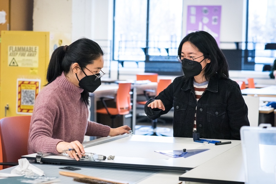 Two people sit at a drafting table in a well-lit studio space.