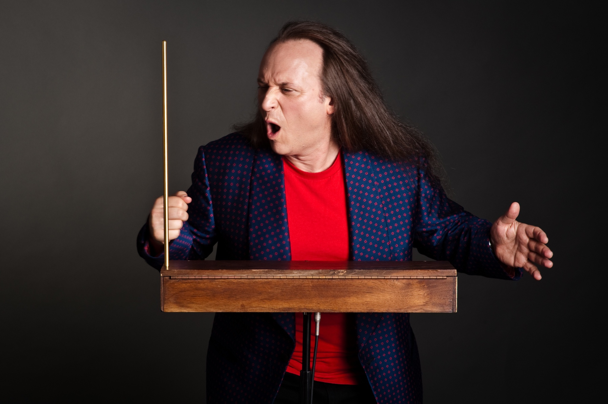 A white man wiith long brown hair stand with arms outstretched behind a theremin. He wears a purple blazer with a bright red tshirt beneath it.