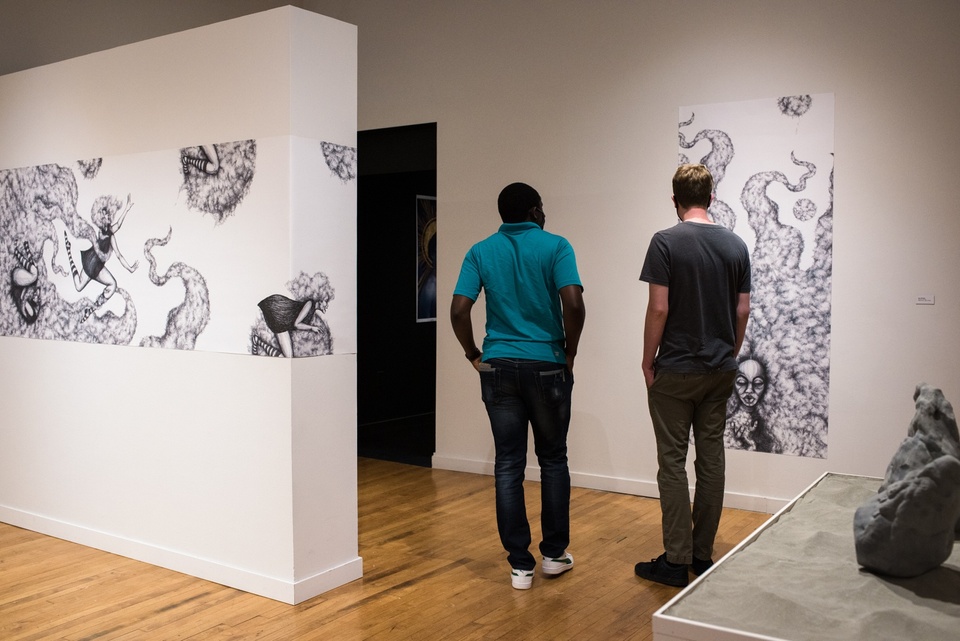 Two people round the corner of a small gallery space towards a black box room. Black and white drawings are on the walls.