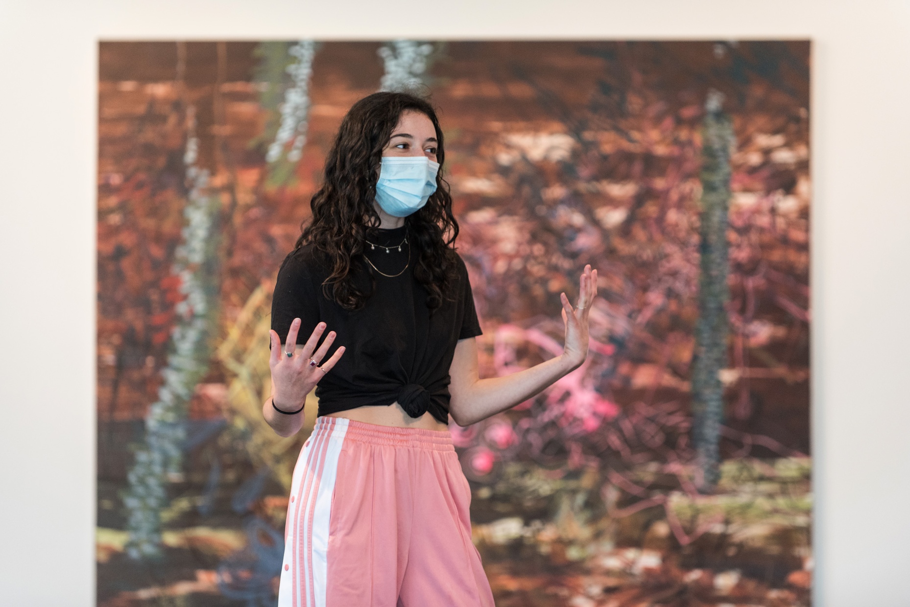 A young woman wearing a mask, a black shirt, and pink pants, stands in front of an abstract painting while speaking and gesturing with her hands.
