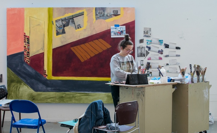 Person works at a standing table full of painting supplies. Behind them is an in-progress painting taking up about 6' of wall space.