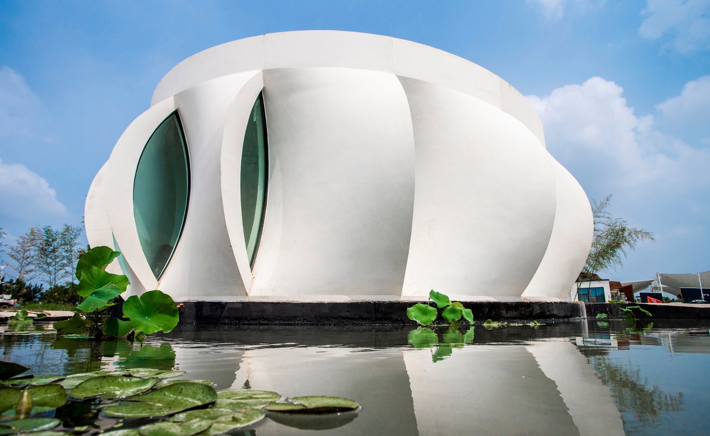 Photo of the exterior facade of a single-story home during the day. The exterior is composed of curved, overlapping panels, arrayed around a central axis like a blossoming flower. Water and lily pads are in the foreground, in front of the home.