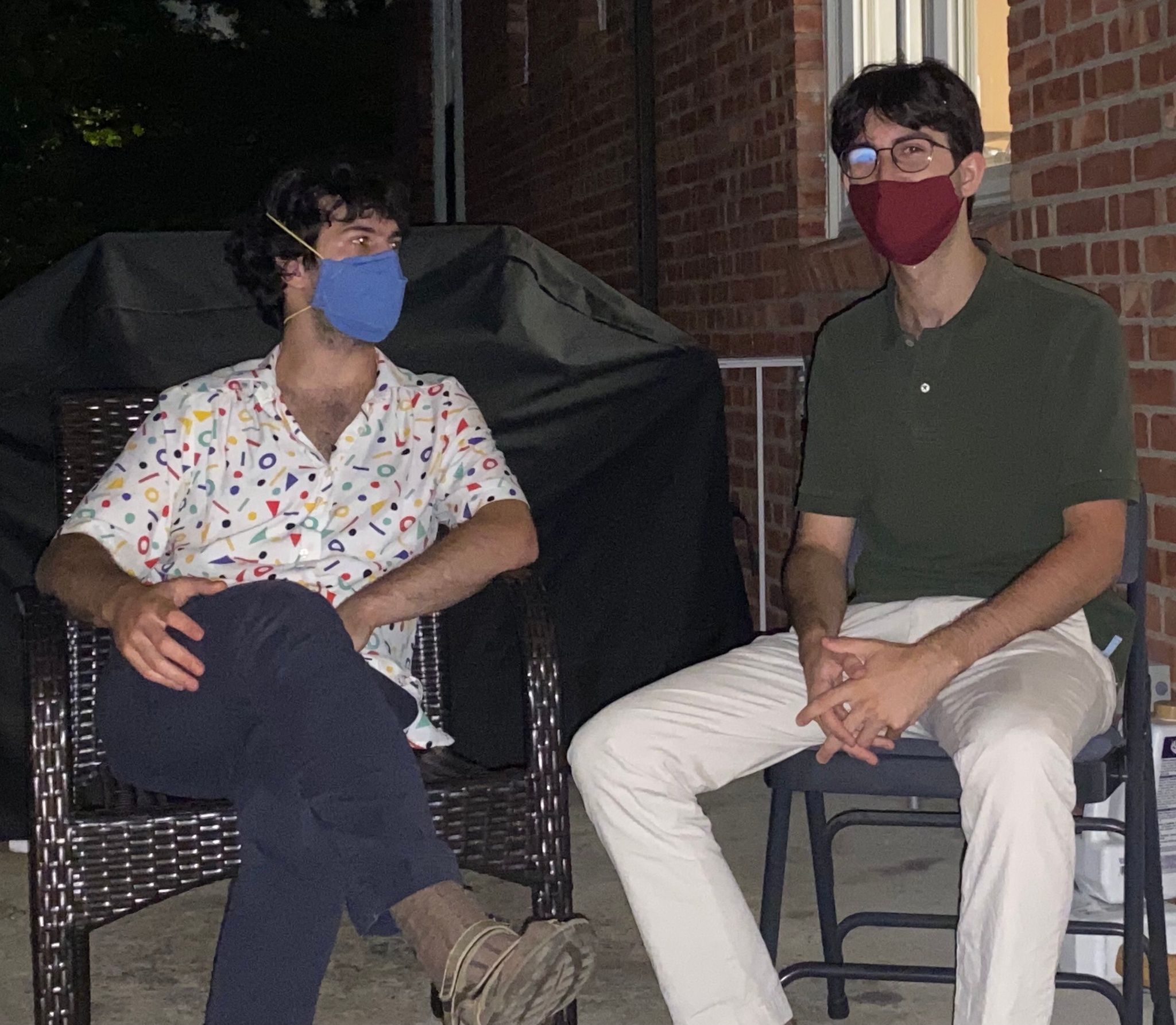 The Brothers Sick: Ezra on the left, Noah on the right. Two white Ashkenazi Jewish disabled brothers with dark brown hair sitting on a porch off of a red brick home on a summer night, dark sky in the background. Ezra is wearing a blue face mask, short sleeve white shirt with red, green, blue, yellow geometric shapes with blue pants, legs crossed and looking at Noah. Noah is wearing a red face mask, round metal glasses, a green short sleeve polo shirt, white pants with hands crossed in lap and legs spread, head looking straight towards the camera. 