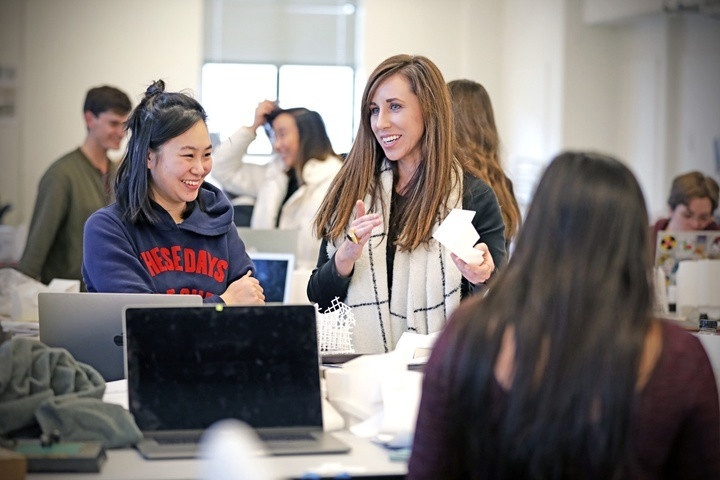 In a light-filled architecture studio, students chat together while working on laptops. At center, a faculty member and student are smiling and engaged in lively conversation.