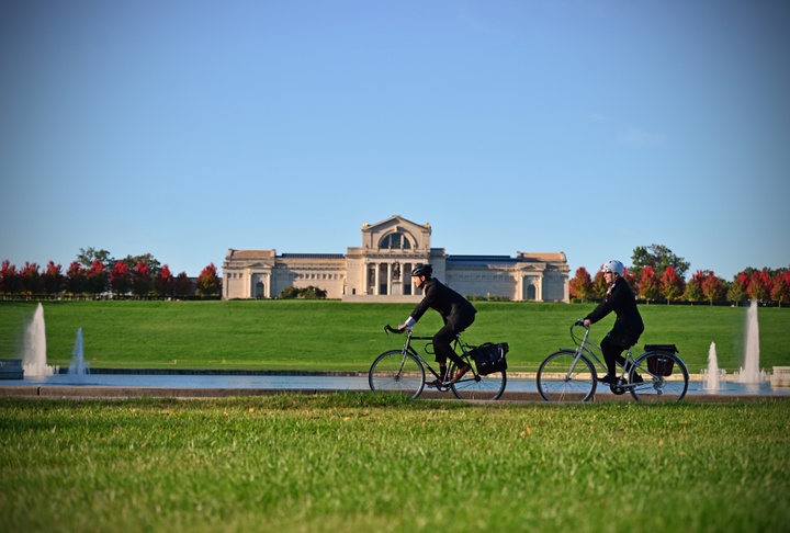 Two people ride bikes along the edge of a pond with fountains. At the top of the hill behind is the Saint Louis Art Museum.