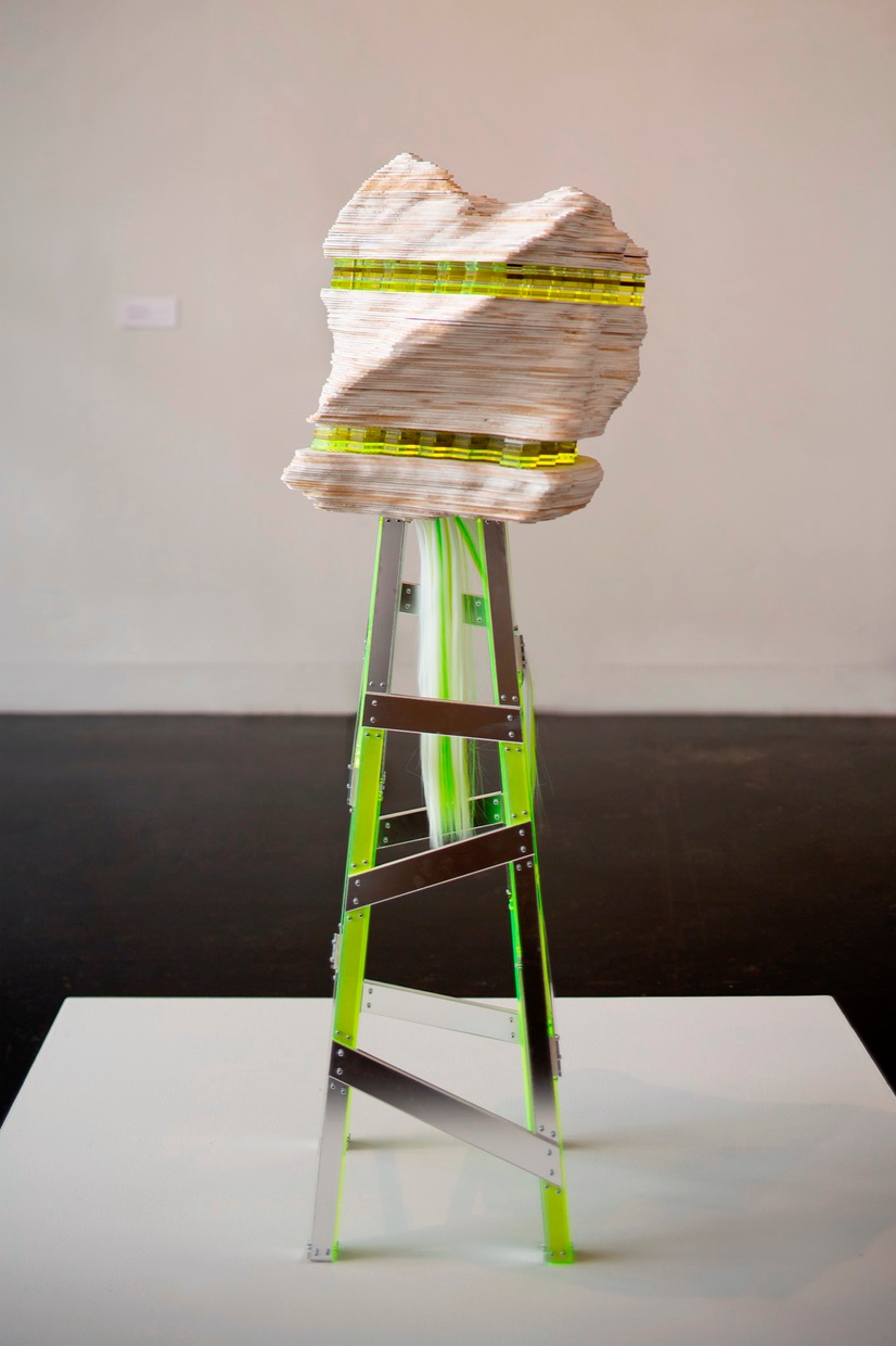 Sitting on a white pedestal is a neon green, silver, and light brown sculpture featuring a stand holding up an organic wooden shape.
