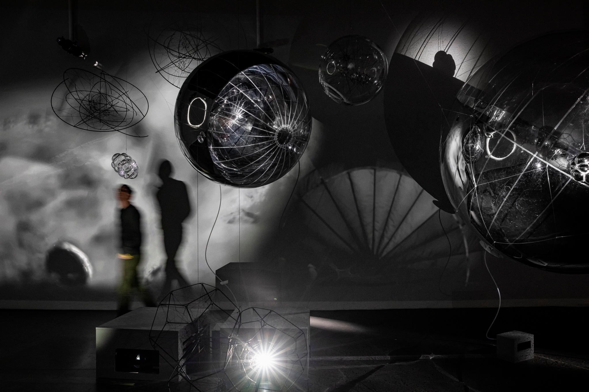 A person walks through a shadowy gallery space, among the play of shadows and reflections cast by large spherical sculptural objects hanging in the space