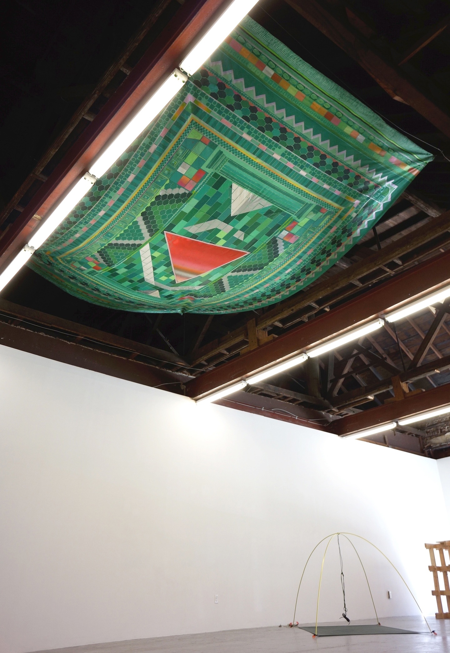 View of a textile hung above among the wooden rafters/joists of a ceiling; along these beams are fluorescent lights which illuminate the gallery space below — the walls are white and there is a wire sculpture below, on the ground in the distance. The textile up above is green and red with a geometric pattern, a (red) triangle in the center.