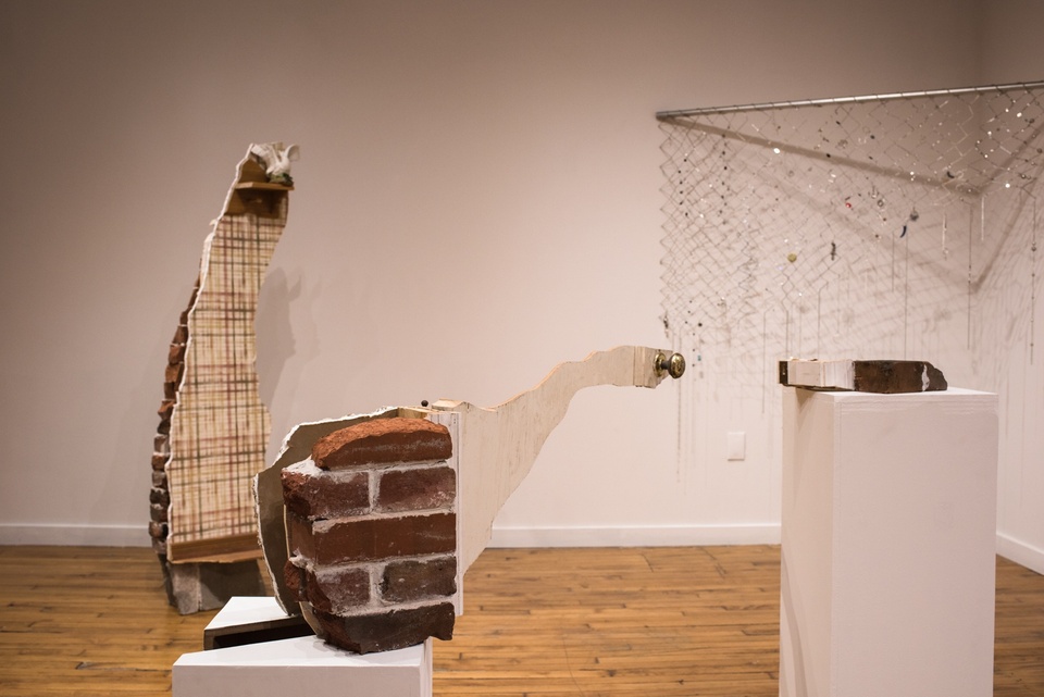 An array of pieces from a brick house are displayed in a gallery space. In back is a tall narrow segment of a brick wall, plastered and wallpapered on one side with a little wooden shelf displaying a headless ceramic goose; a piece of brick wall with a little sliver of a white wooden door including a door handle sits on a plinth; another wooden segment is on a plinth opposite. On the back wall is a metal bar with dangling chain link covered with gems and trinkets.
