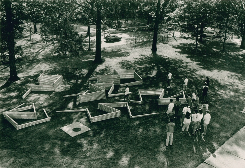 Black and white photo - overhead view of people on a lawn assembling a maze-like structure out of wooden ties.