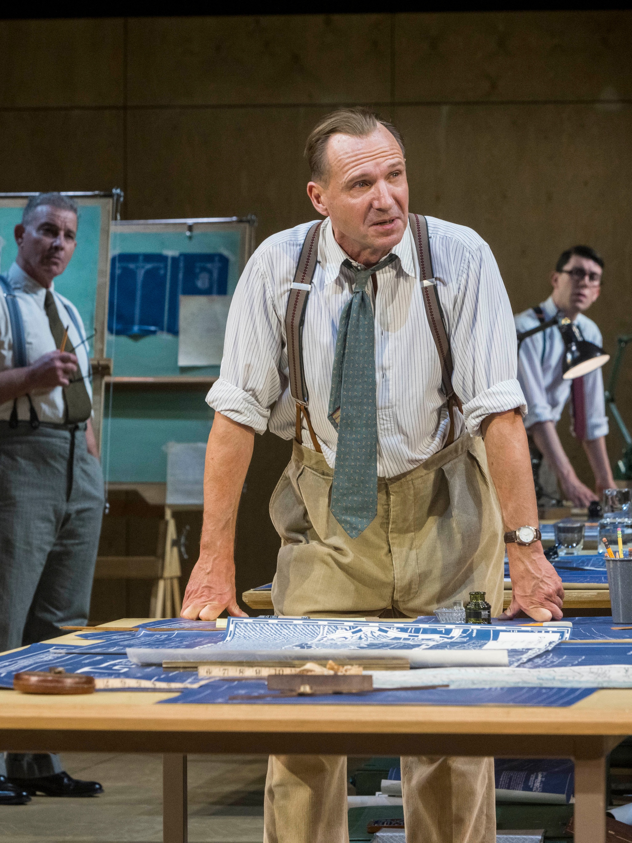 The actor Ralph Fiennes, a white man, stands leaning with both hands on a draftsman's table. He is in costume as Robert Moses, wearing khaki pants, suspenders, and a blue gray blue tie. In the background are two other actors dressed similarly looking toward him.  