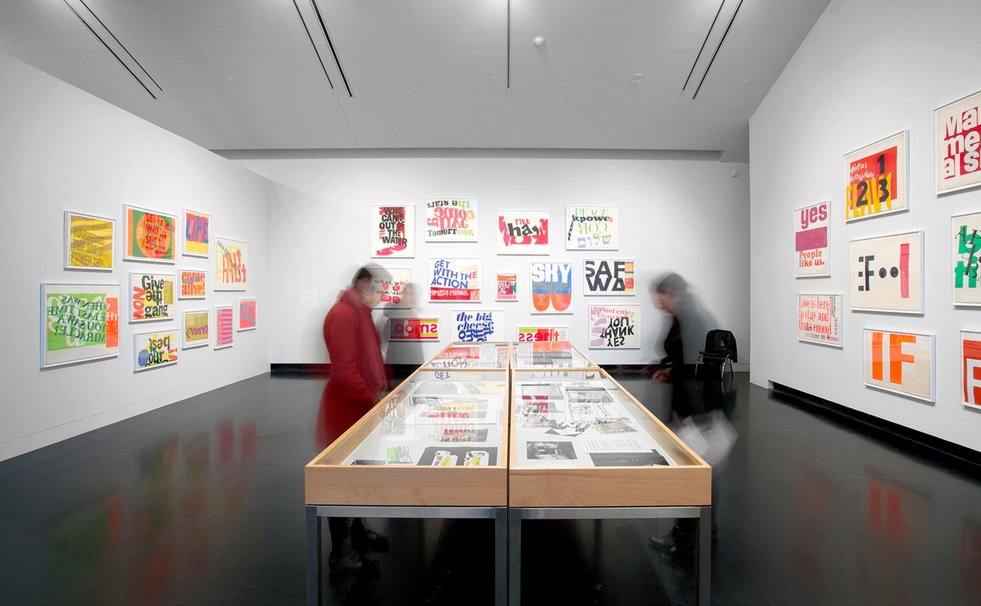 Two wood and glass cases with screenprints inside in the middle of a gallery space with white walls and colorful prints hung on the walls.