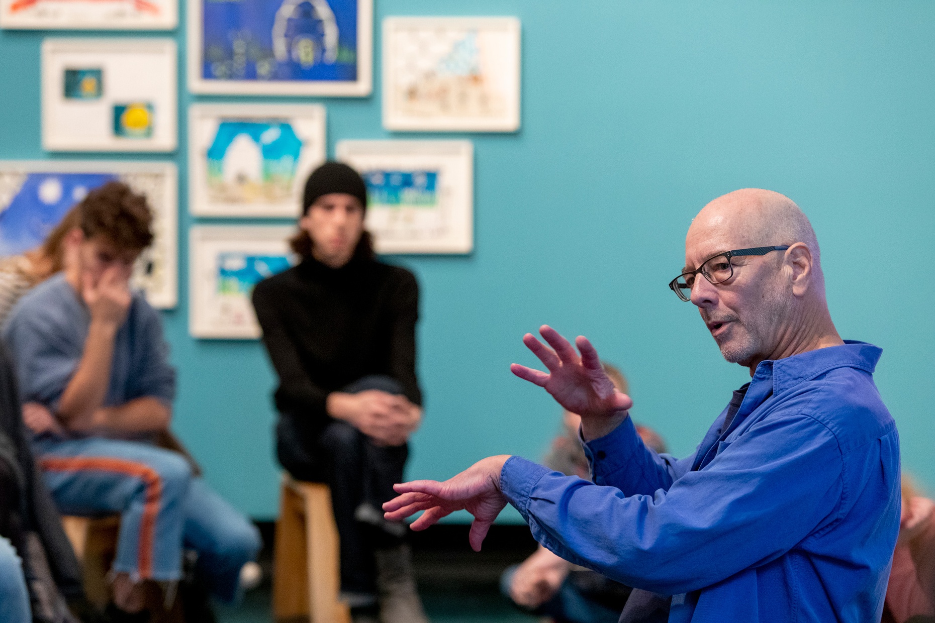 A white, older male talks to a group of people in front of a blue wall.