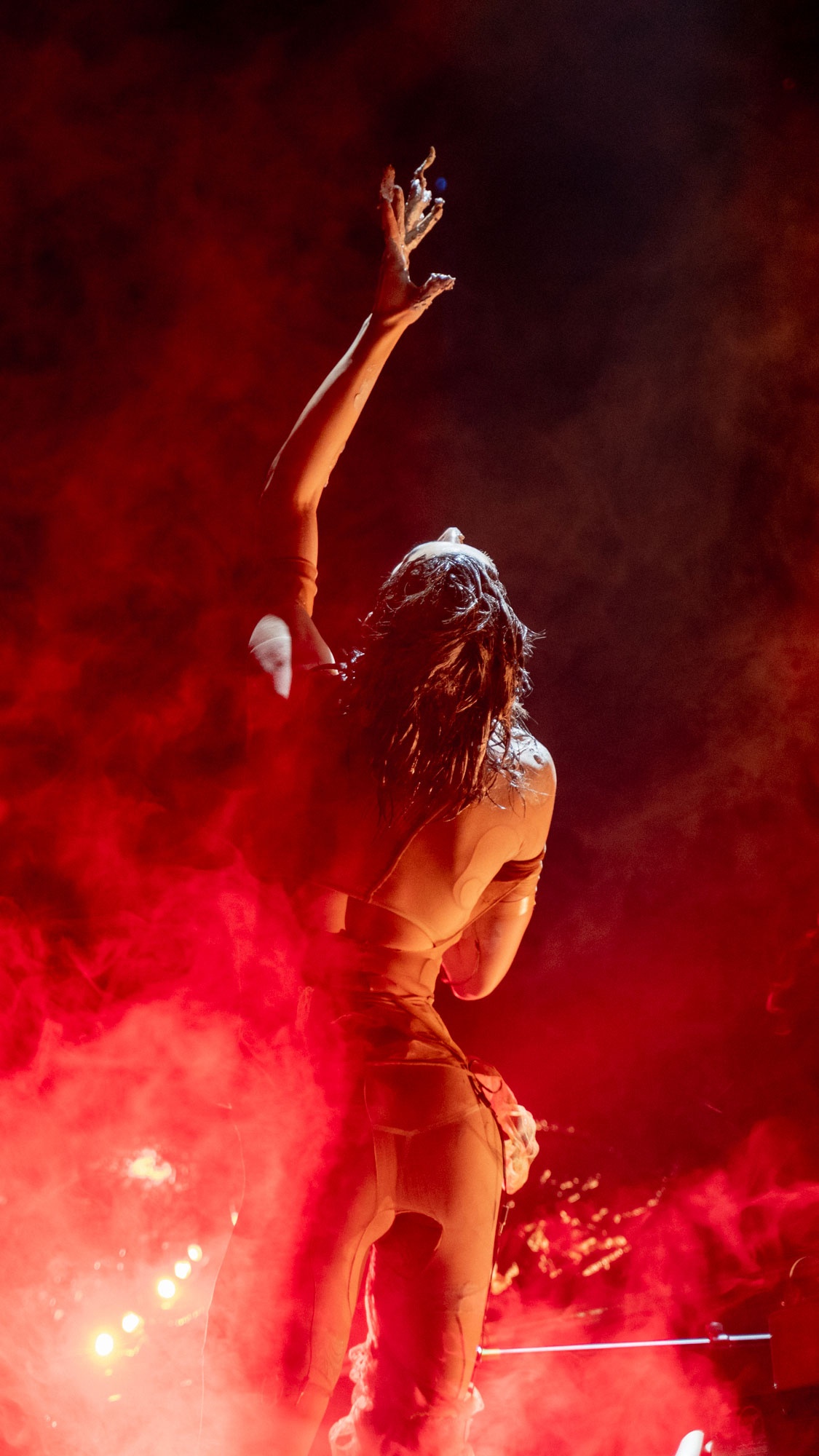 Arca performing on stage with arm in the air and red smoke.