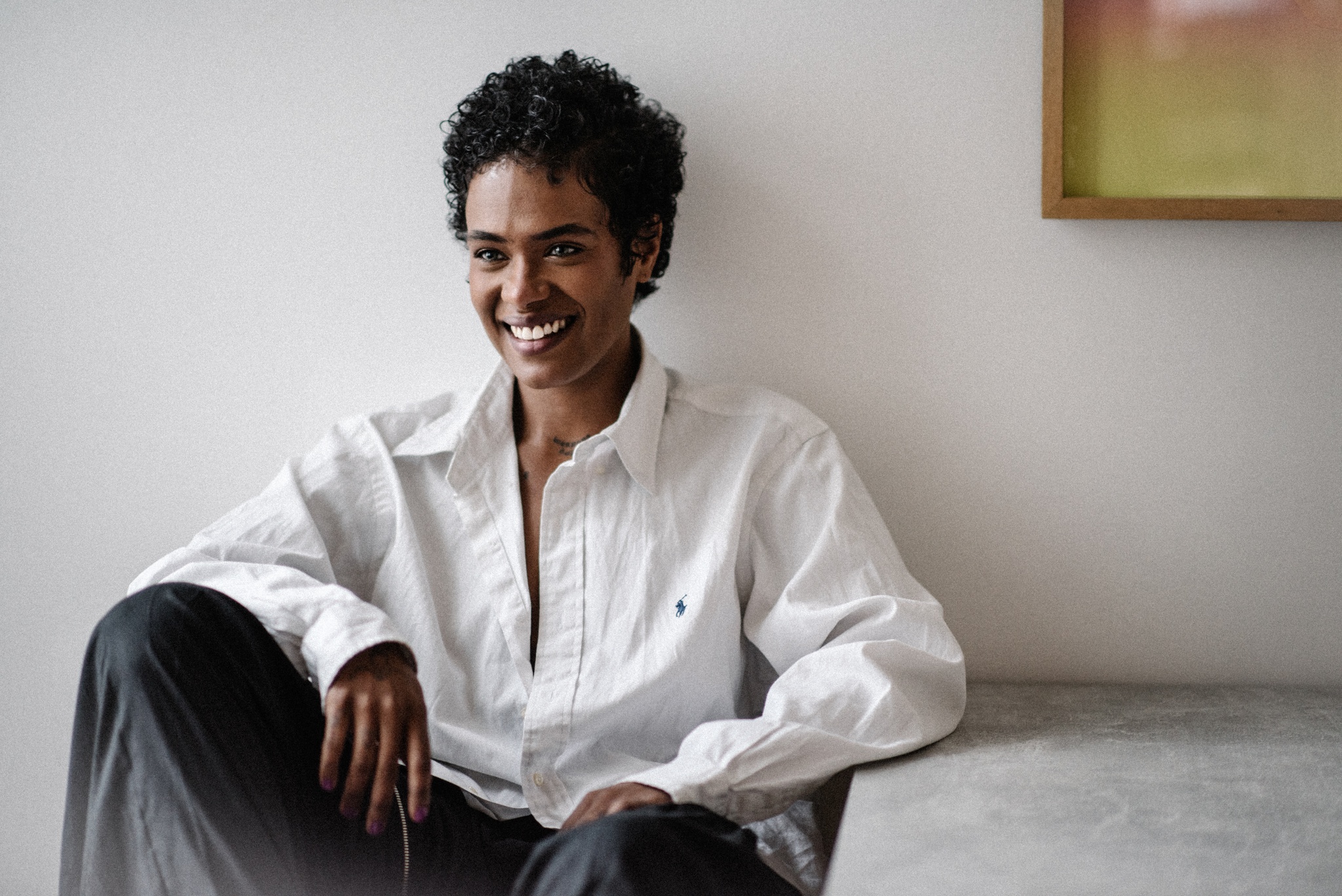 A portrait of Navild Acosta, an Afro-Latinx queer trans person who poses sitting against a white wall. He wears an oversized white button down shirt and sits with one leg pulled up toward him. He smile broadly and props one arm on a table and the other on his knee. 
