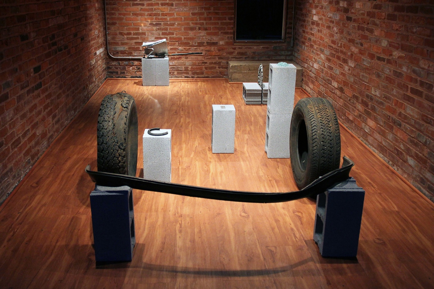 In a gallery space, whose walls are brick with wooden floors, an installation: various car parts placed on cinder blocks, some of which are stacked. Two tires are placed opposite each other, in the midground.