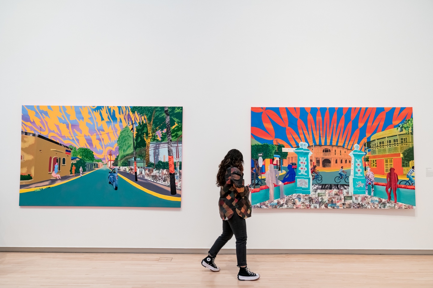 A visitor observes two large brightly colored and patterned paintings. The one on the left depict a narrow roadside and the one on the right depicts a town square.