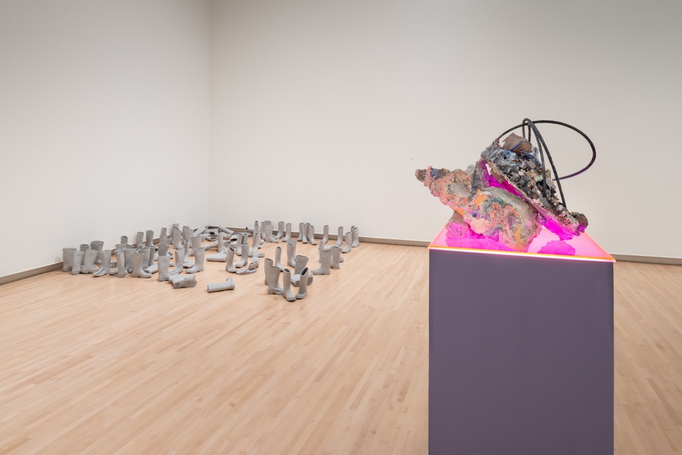 Overview of gallery with one sculptural object made of various materials on a plinth lit from below by neon pink light, and a large arrangement of grey rubber boots on the floor.