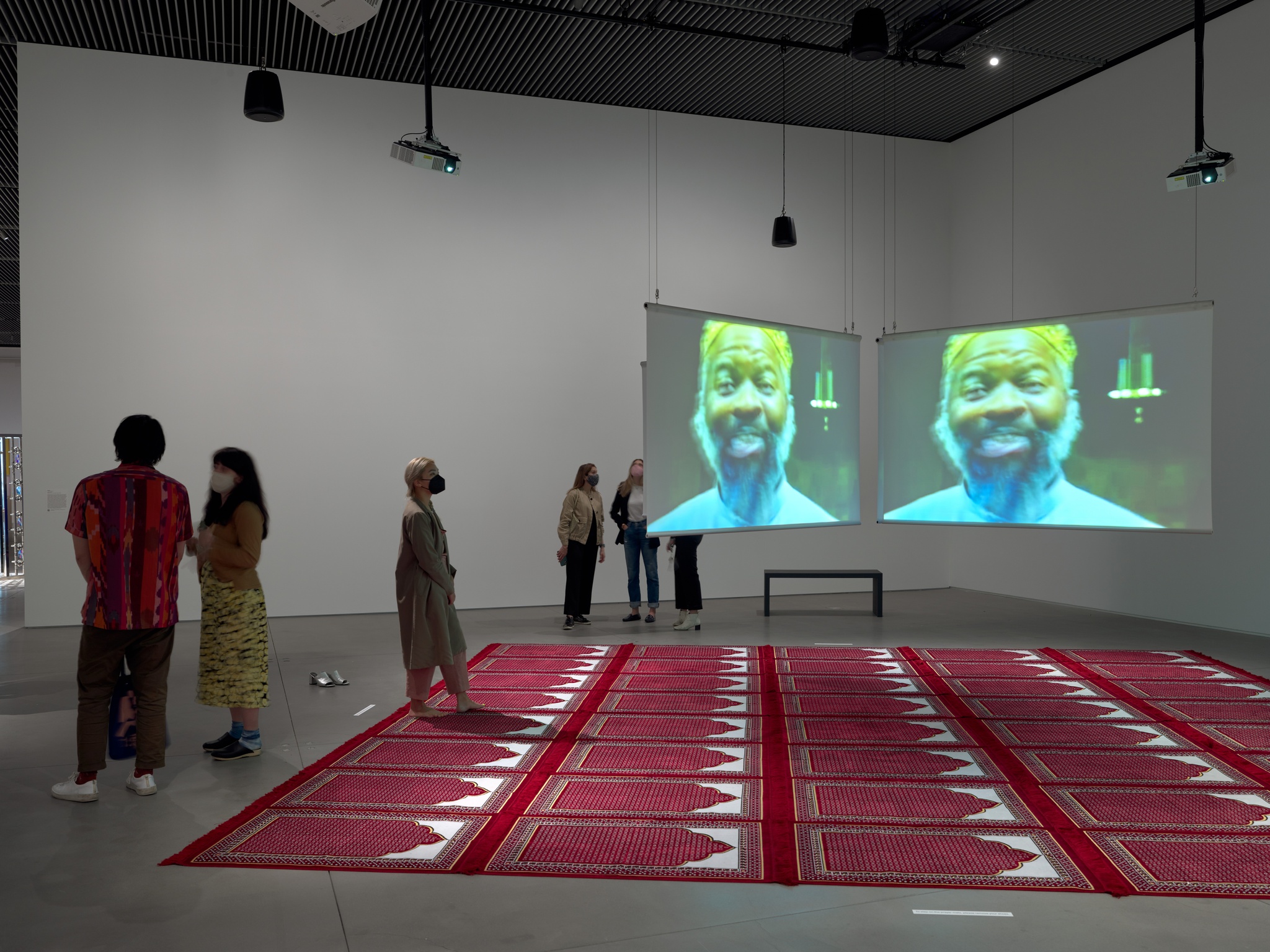 An installation in an art gallery comprised of red prayer rugs creating a rectangular space on the floor with two screens hanging over them with the same image of a man's face projected on them. Some people stand to the left of the rugs and one person steps onto them.