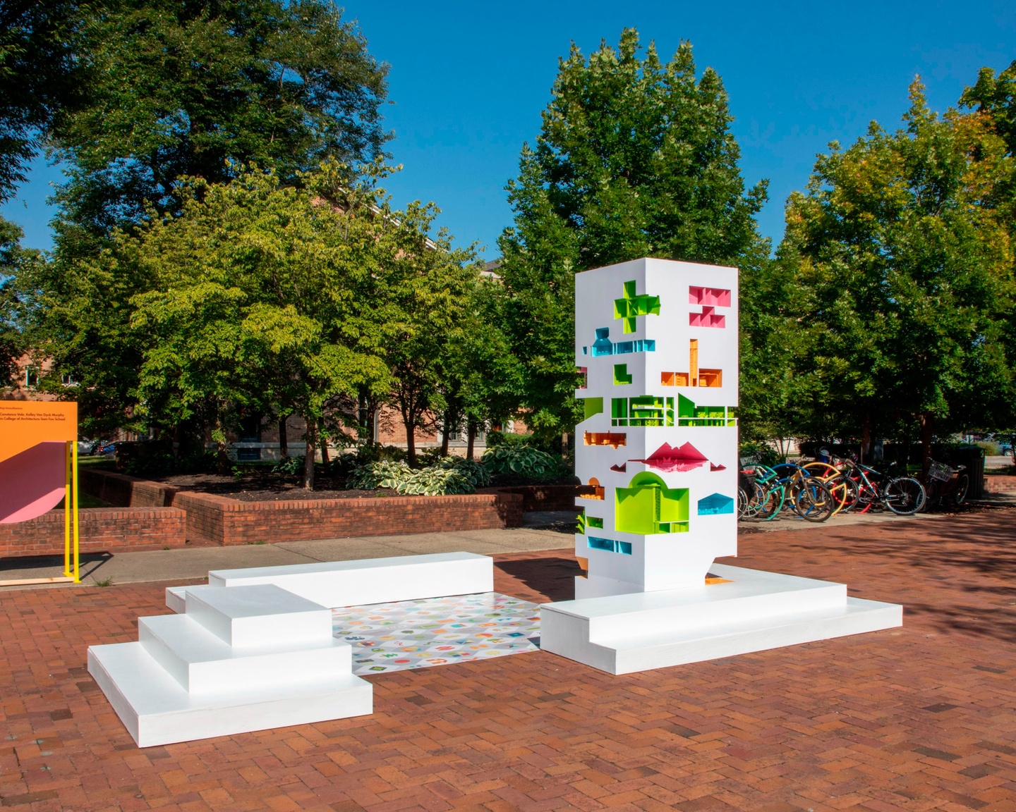 large white dollhouse with colorful sections cut into it with dolls made by students sitting outside in a plaza on brick with surroundings trees