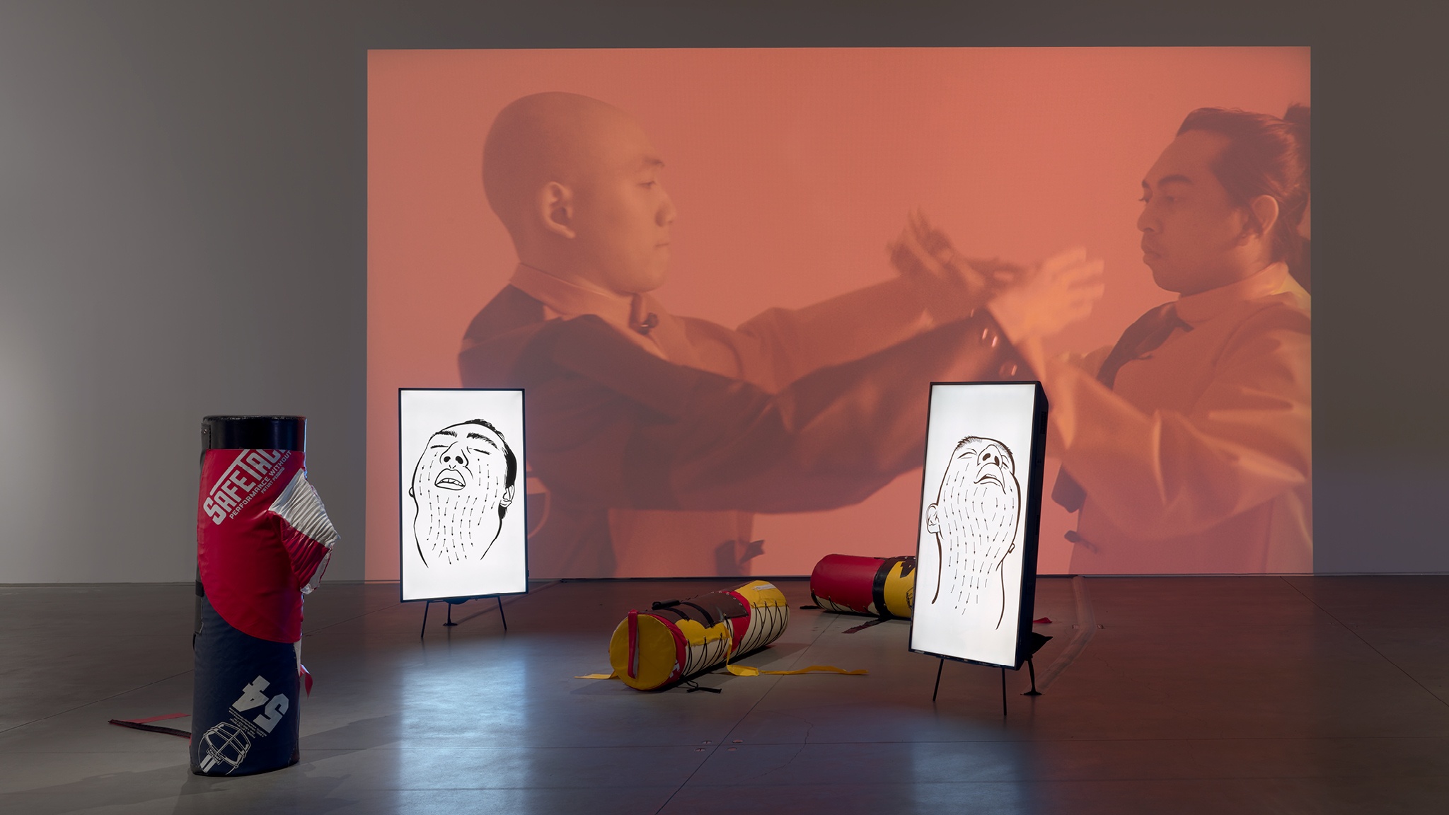A video projection of two men with their arms outstretched toward each other on a wall with deconstructed tackling dummies and two vertical video monitors on the floor in front of the projection. The video monitors both show drawings of men's faces turned upward. 