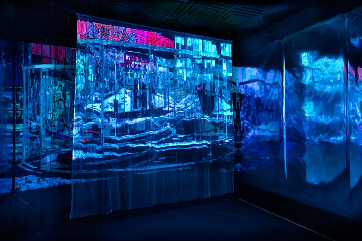 Black box room with a bright blue projection of an impressionistic cityscape against several semi-transparent curtain panels.