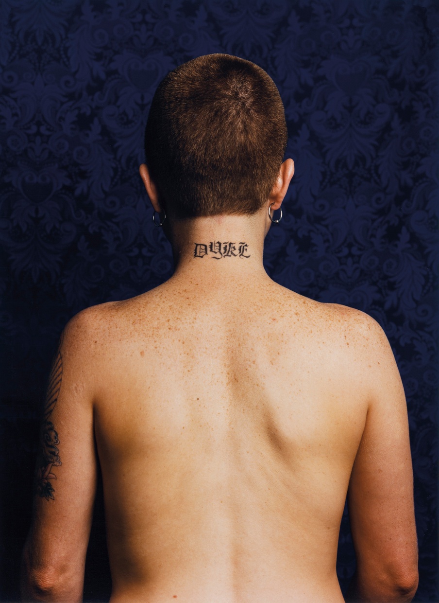 A photograph of the back of a nude, light-skinned female figure with short dark hair and the word “Dyke” tattooed on their neck.