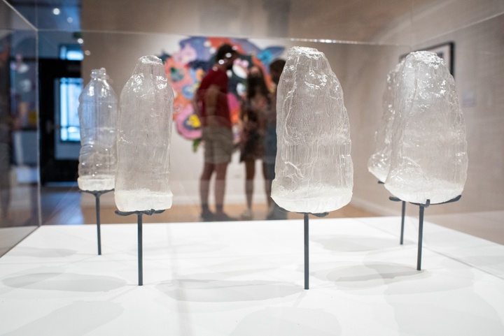 Close up of five clear stone-shaped objects affixed to prongs over the top of a white plinth. Behind these is a gallery space with several visitors.
