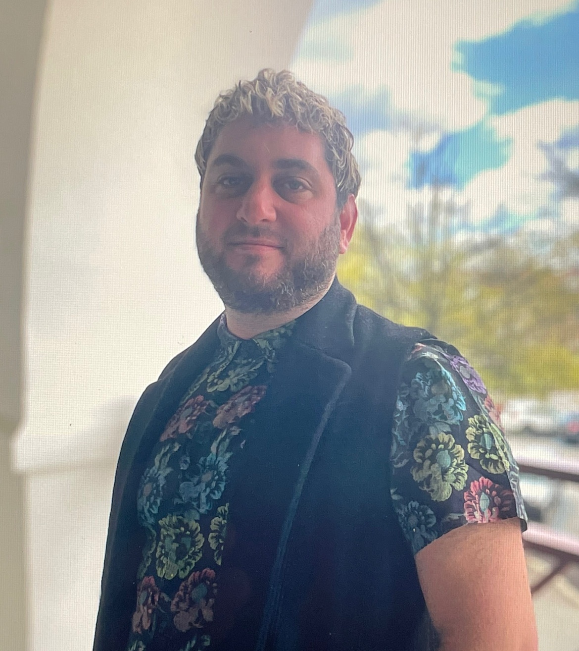 A photo of Jesse Bandler Firestone posing against an outdoor archway with a blue sky and trees in the background. Jesse wears a vest over a short sleeve shirt with a blue, green, and pink floral print. 