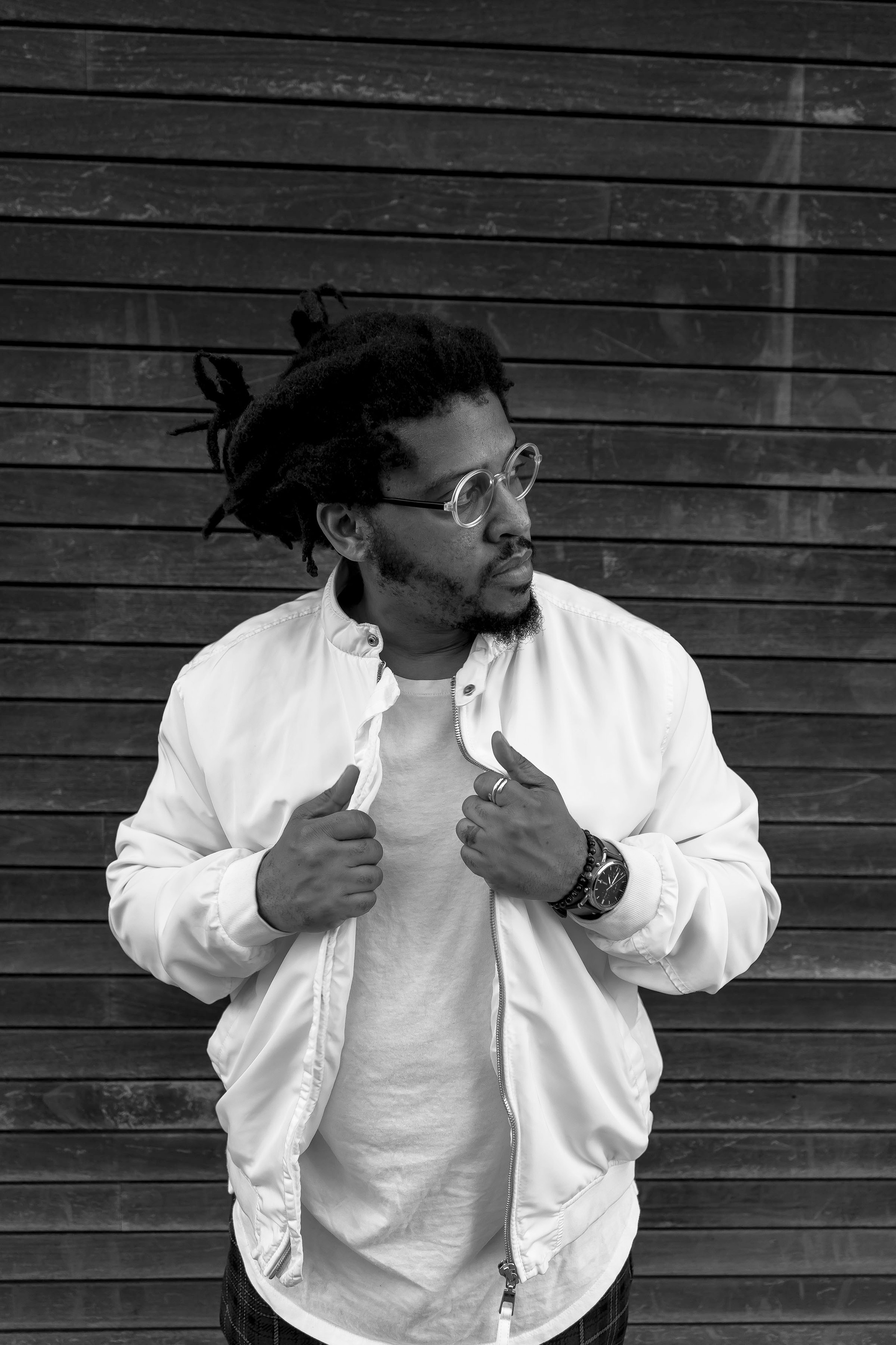 A black-and-white photo of artist Kamau Ware standing against a wooden wall. Ware wears translucent-framed glasses, has his hair pulled up behind his head, and has both hands pulling on the lapels of a white bomber-style jacket while he looks away to his left. 