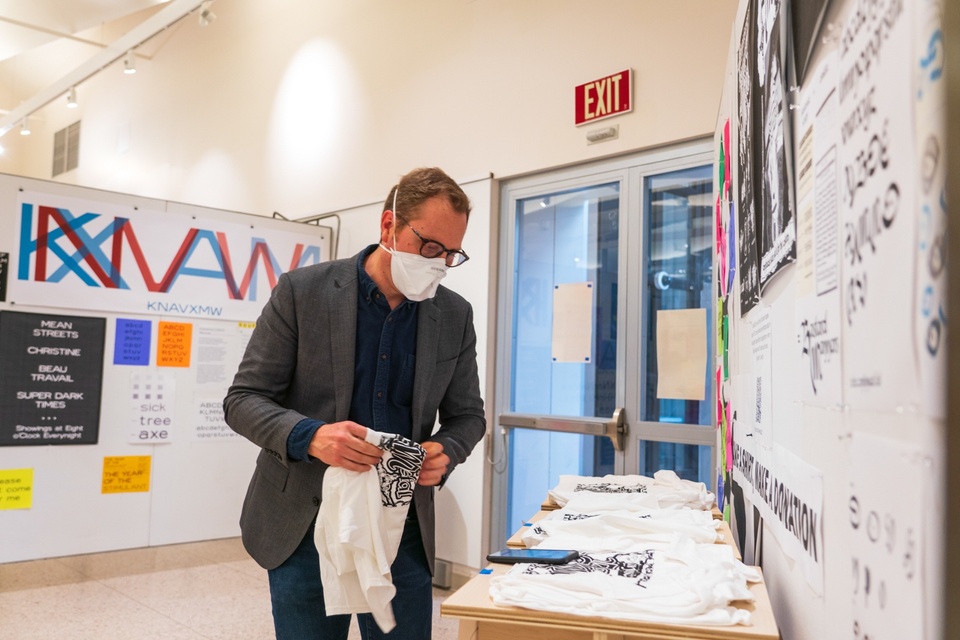 Faculty inspects a table of white tshirts with a black imprint on them, placed in front of a wall of blackletter typography samples.