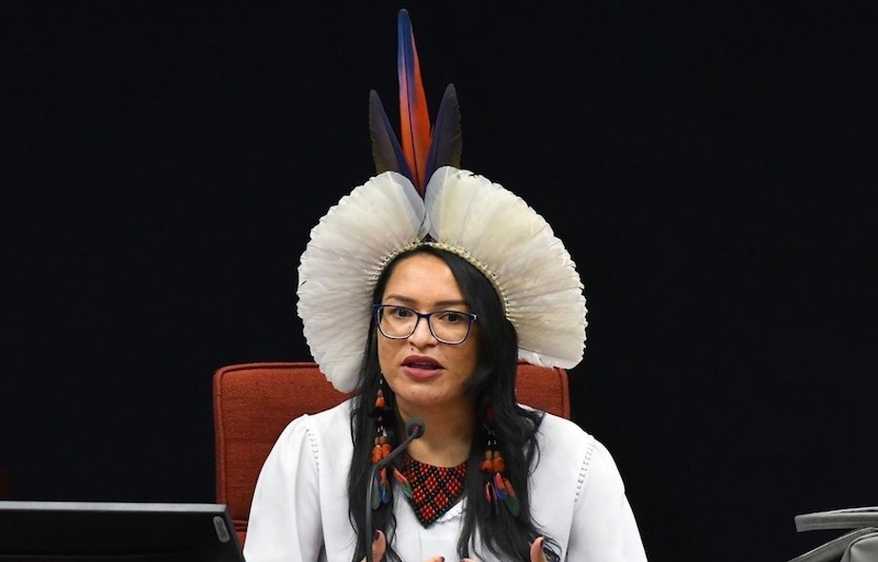 Samara Pataxo, an Indigenous woman from Brazil, sits in front of a microphone. She has long dark hair and wears a headdress of white feathers. A the center of the headdress are three blue and red feathers that stand straight up above the white feathers. Samara speaks into the microphone.