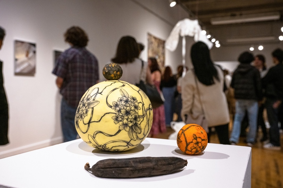 Closeup of an artwork on a pedestal in a gallery space. Four pieces of fruit are arranged together - a melon, an orange, a desiccated clementine, and a very brown banana. The melon and orange have intricate black linework tattoos of flowers twining around them. A flower pattern has been carved into the clementine, and the banana is just very dark all over.