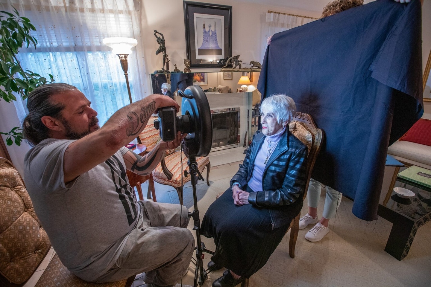 A man takes a photo of a woman, who is seated against a black backdrop.