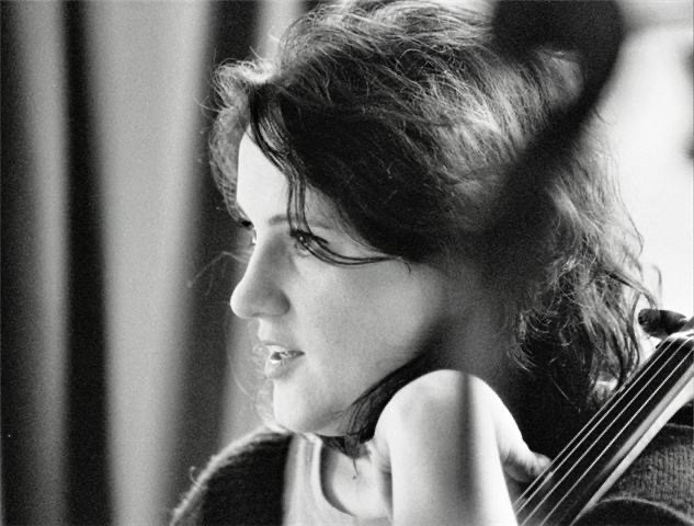 A woman seen in profile from the shoulders up. She cradles the neck of a cello between her upheld arm and shoulder. 