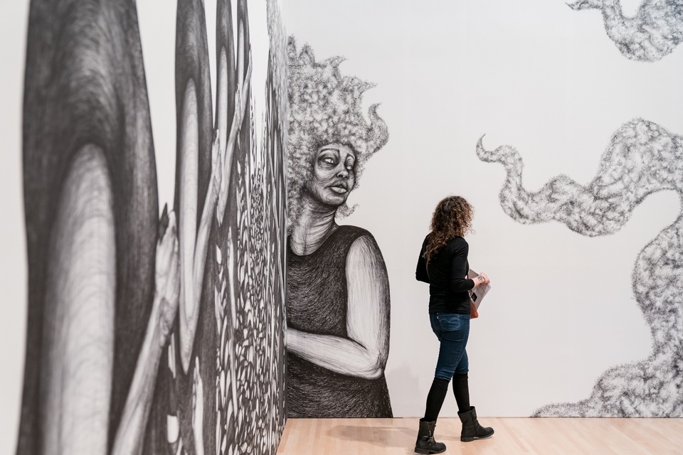 A person walks past a white wall with a large-scale black and white drawing of women in black dresses with pouffy hair. More disembodied hair trails in from the right side of the frame.