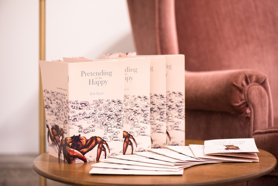 Close shot of several brochures piled on an end table next to an armchair entitled "Pretending to be Happy" with a drawing of a crab on a beach.
