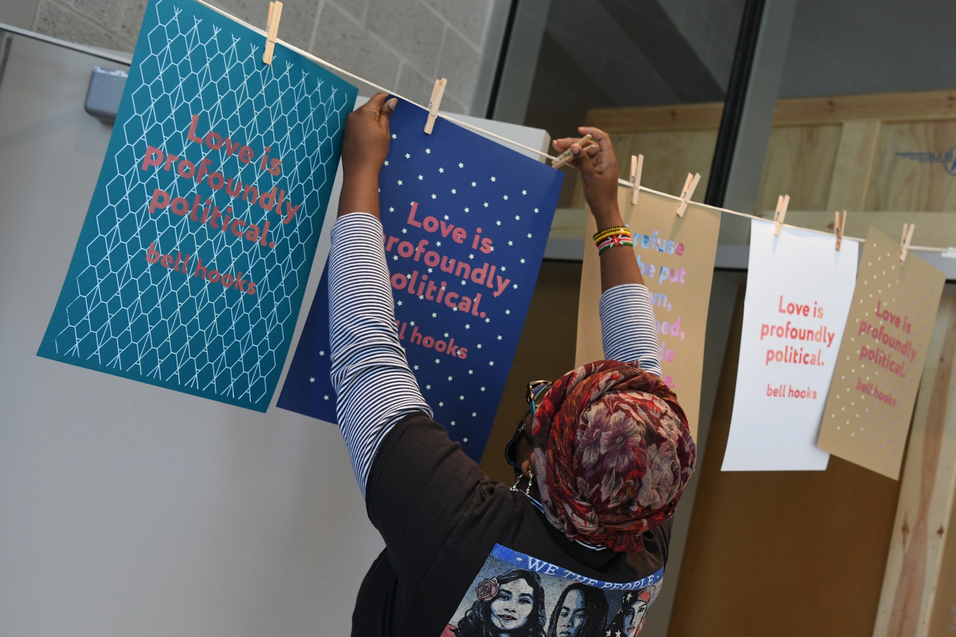 A student, with her back to us, reaches up to hang a poster to dry on a clothesline. The poster most visible reads "Love is profoundly political. bell hooks".